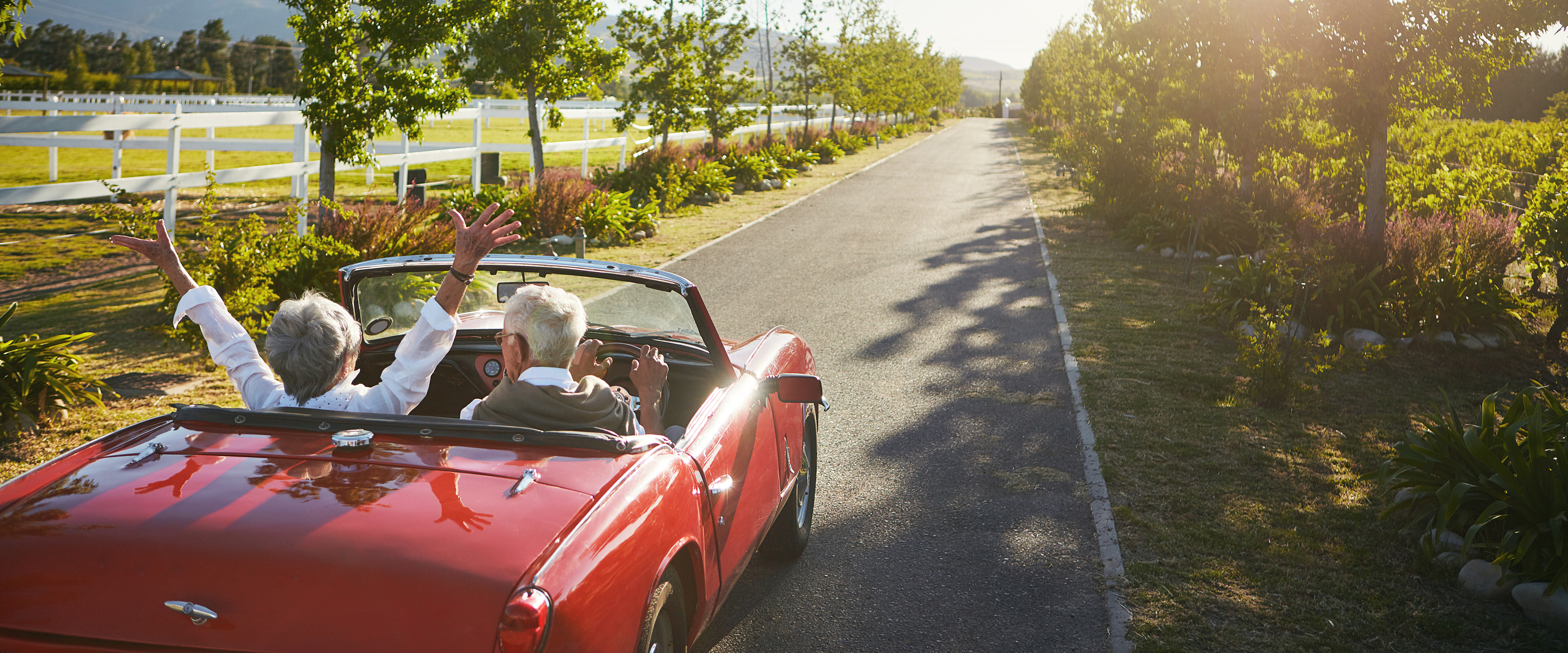 A senior couple drives in a red car with the top down. The woman throws her hands in the air.